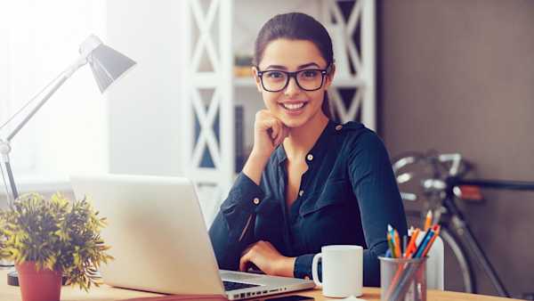 woman with glasses and coffee looks at camera in front of open laptop