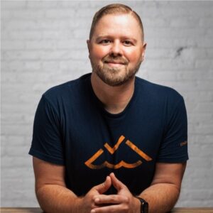 Portrait of a smiling man with short blond hair and a beard, wearing a navy T-shirt with an orange logo, seated at a table against a light brick wall.
