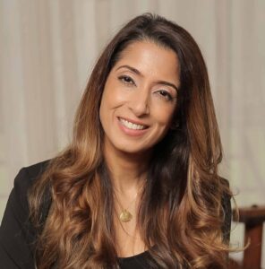 3B9A8476 - Sonia Bhandari - Coach Training EDU Smiling woman with long wavy brown hair in a black top, seated indoors against a neutral background.