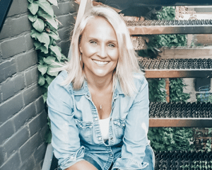 Smiling woman with blonde hair wearing a light denim shirt, seated outdoors near green plants