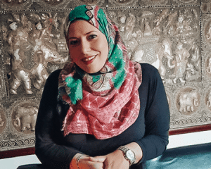 Smiling woman wearing a colorful patterned hijab, seated in front of a decorative metal wall panel