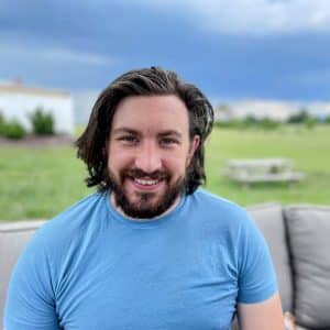 Smiling man with dark wavy hair and a beard wearing a light blue t-shirt outdoors on a patio