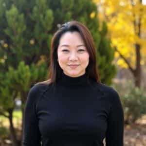 Smiling woman in a black turtleneck posing outdoors with fall foliage in the background.