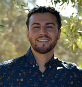 Smiling man with dark hair and a beard wearing a navy floral button-up shirt outdoors with sunlit foliage in the background