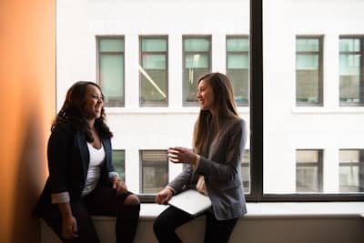 two women sit on a windowsill of a large building, holding a conversation