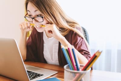 a woman in front of a laptop, biting a pencil with a stressed expression