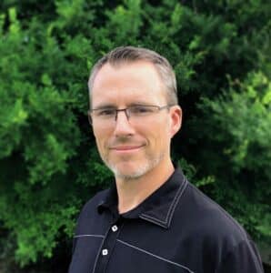 Head-and-shoulders portrait of a man with short gray-streaked hair and glasses, wearing a black shirt, outdoors with green foliage behind him.