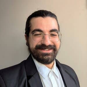 Smiling man with dark hair and beard, wearing round glasses and a black blazer over a light shirt, against a neutral wall background.