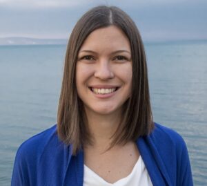 Csontos Timi headshot - Coach Training EDU Smiling woman with shoulder-length brown hair by the sea, wearing a white top and blue cardigan, with a cloudy sky above.