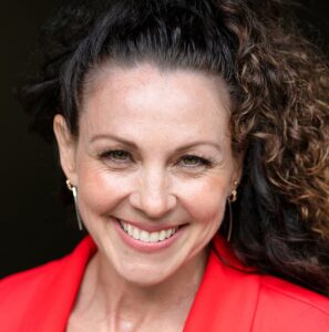 RedSmilesmaller1080 - Coach Training EDU Smiling woman with dark curly hair in a red blazer and black lace top, posing for a portrait.