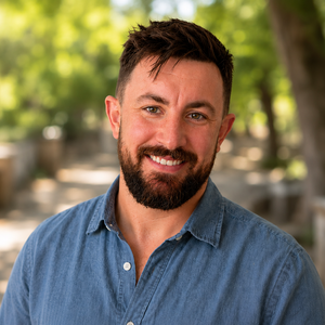 Smiling man with short dark hair and a beard wearing a blue button-up shirt outdoors