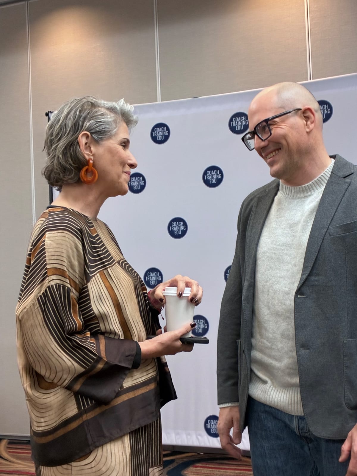 Two attendees smile and talk in front of a Coach Training EDU backdrop