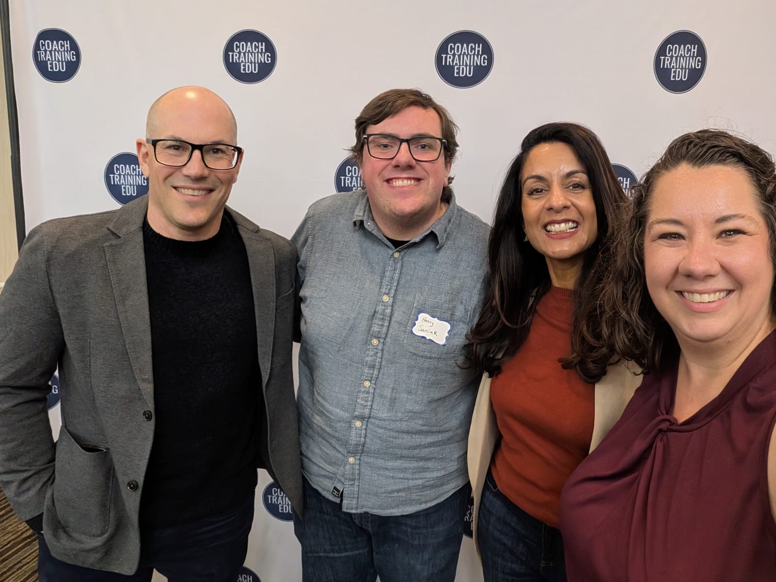 Four attendees smile close together for a selfie in front of a Coach Training EDU backdrop