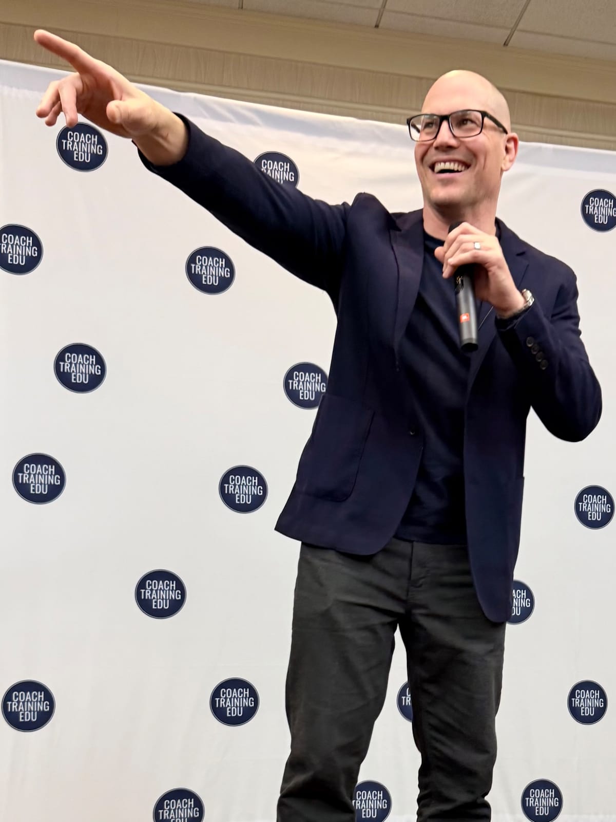 John Andrew Williams, smiling and pointing toward the audience, delivers the opening keynote in front of a Coach Training EDU backdrop