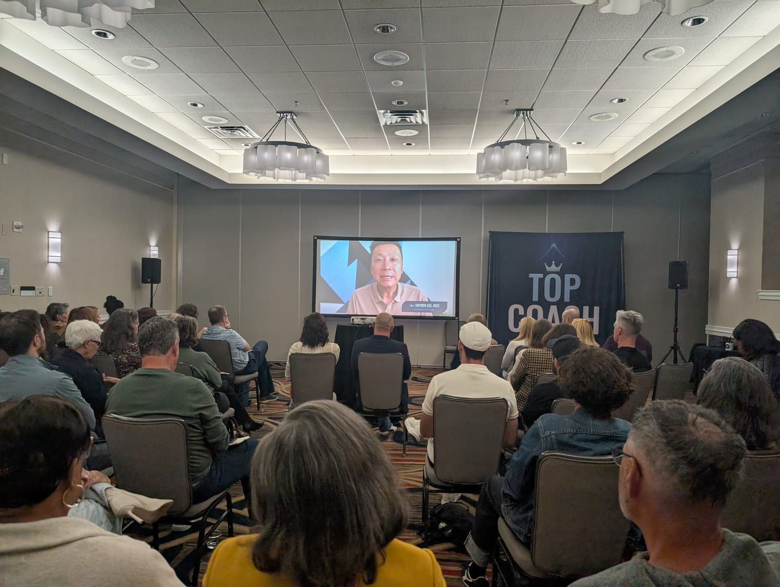 Attendees seated in a ballroom watch a Top Coach video message on a large screen